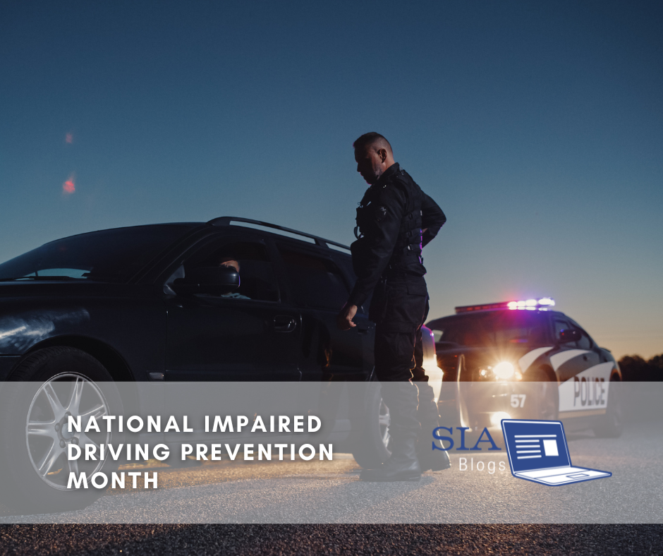 A police officer stands by a car on the side of the road at dusk, with emergency lights flashing on a nearby police vehicle. The scene represents National Impaired Driving Prevention Month.